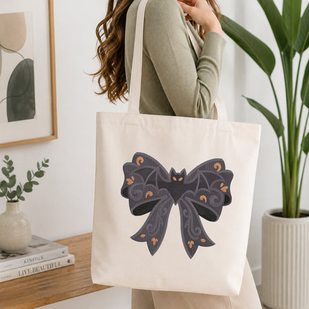 A person wearing a green long-sleeve top and carrying a natural canvas tote bag printed with the gothic bat bow machine embroidery graphic.