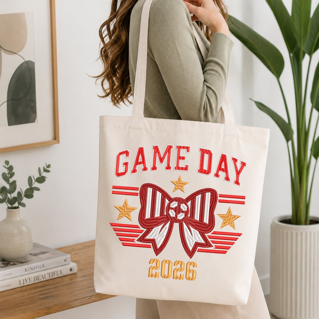 A person carrying a canvas tote bag decorated with the "GAME DAY 2026" embroidery design.