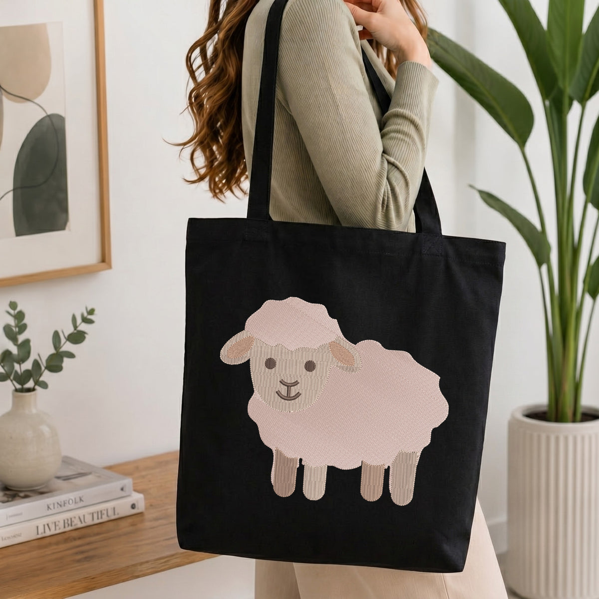 A woman wearing a sage green ribbed sweater carries a black canvas tote bag with a large pink and beige sheep graphic on the front, set in a bright, modern room with a wooden table, books, and a potted plant.