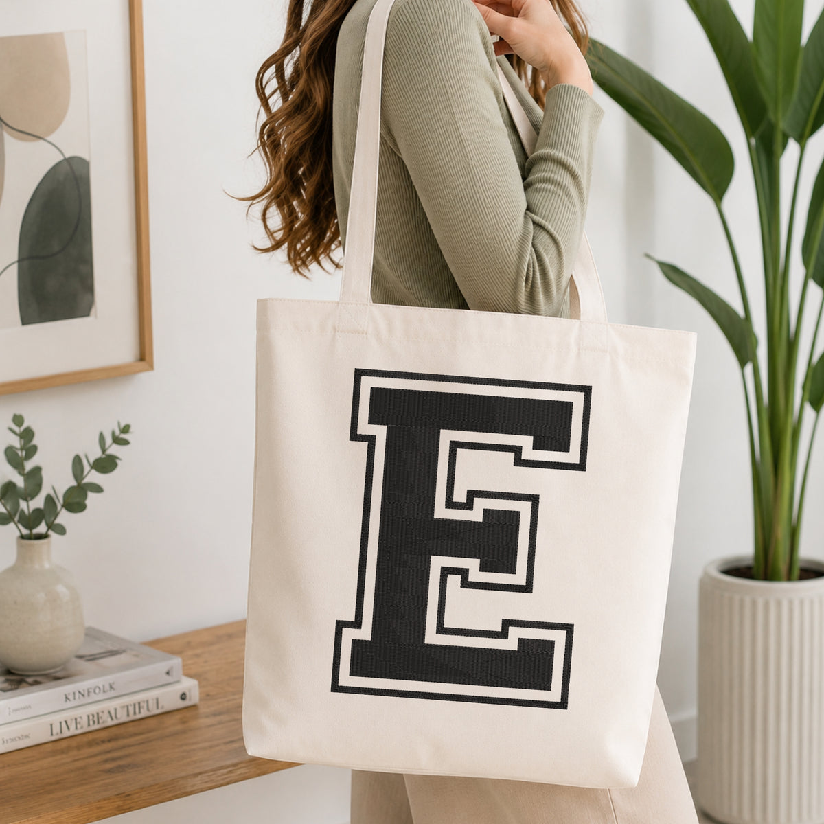 A woman wearing a sage green ribbed long-sleeve top carries a cream canvas tote bag with a large black and white block letter 'E' printed on the front, set in a bright, modern room with a wooden console table and a potted plant.