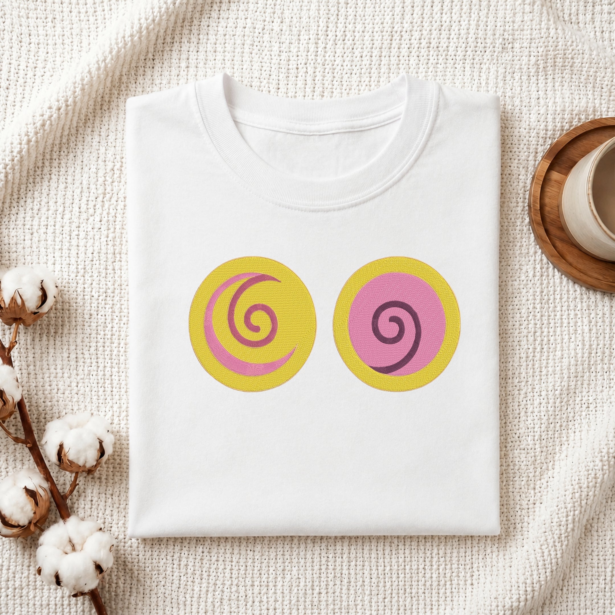A folded white t-shirt with two circular graphic prints on the front, styled on a textured cream knit blanket with cotton bolls and a wooden cup in the background.