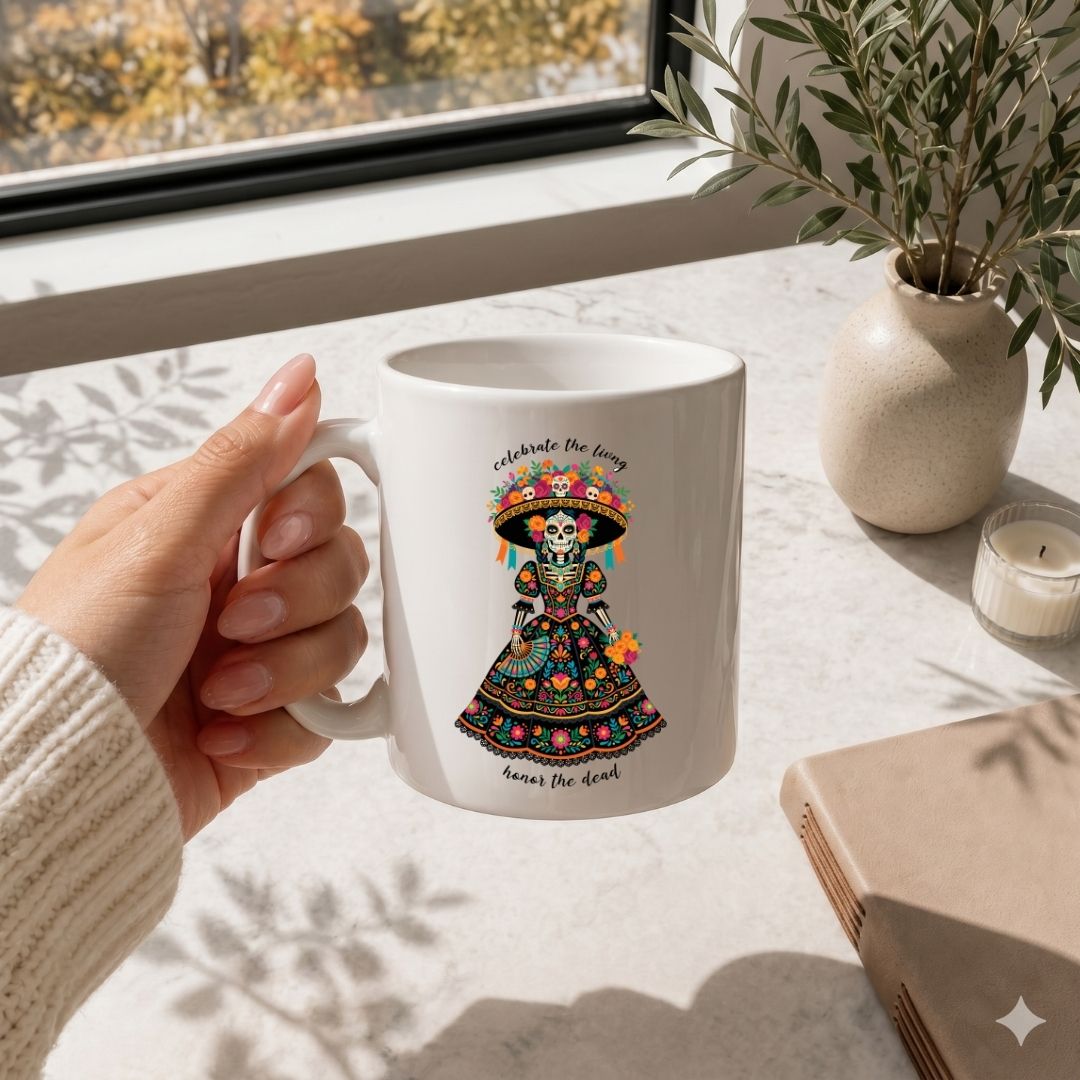 A hand holds a white ceramic mug featuring a colorful Day of the Dead sugar skull illustration with the text 'celebrate the fear' and 'honor the dark'. The mug is shown in a bright, sunlit room on a marble-like surface next to a potted plant, candle, and book.