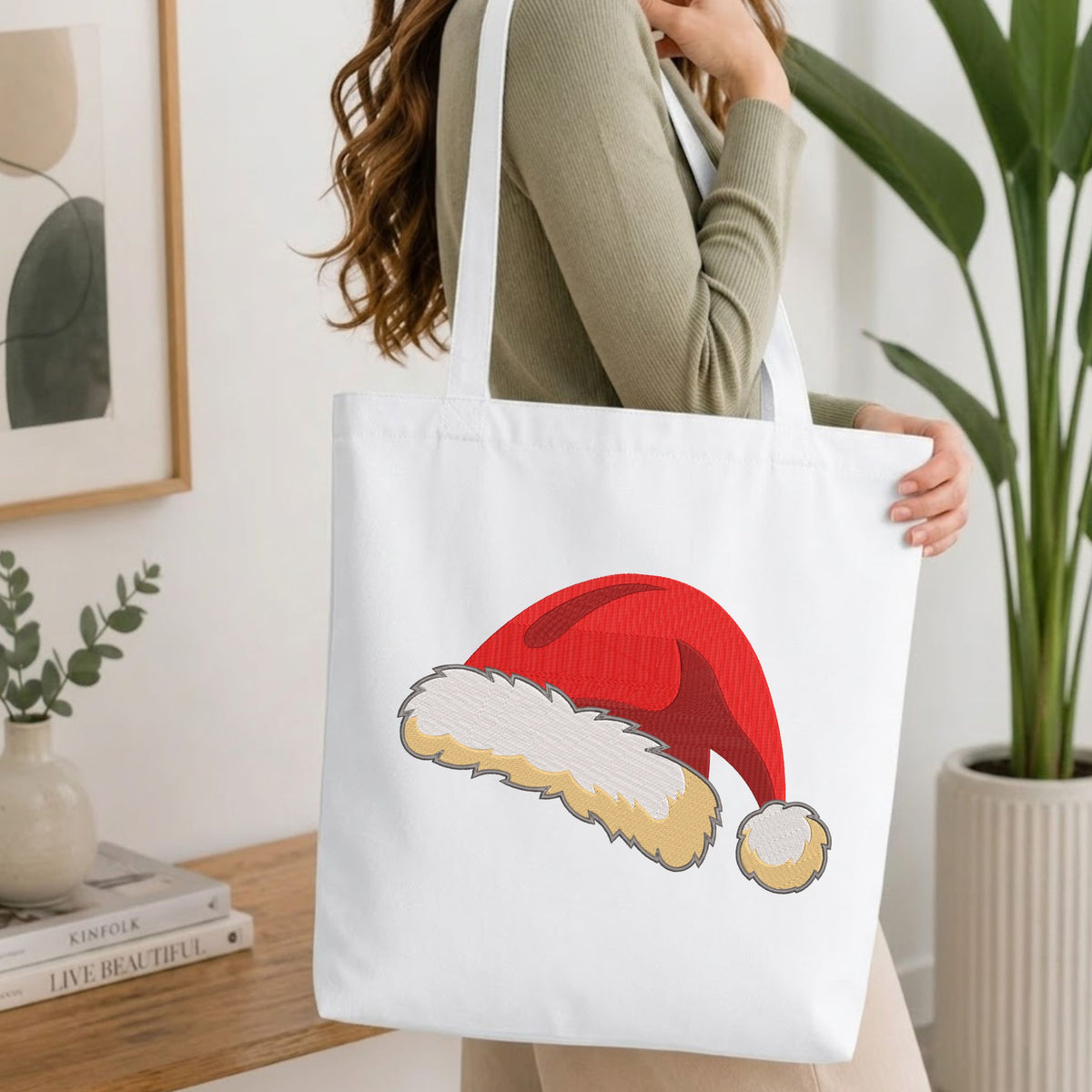 Woman carrying a natural canvas tote bag featuring an embroidered Santa hat design in a bright, modern interior setting.