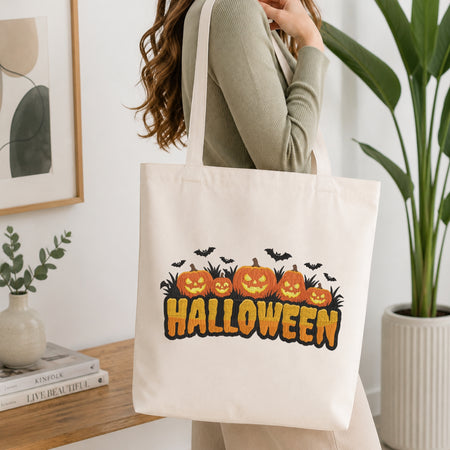 A woman wearing a sage green ribbed long-sleeve top carries a cream canvas tote bag with a Halloween graphic. The graphic features five orange jack-o'-lanterns, black bats, and the word 'HALLOWEEN' in orange with black outlines.