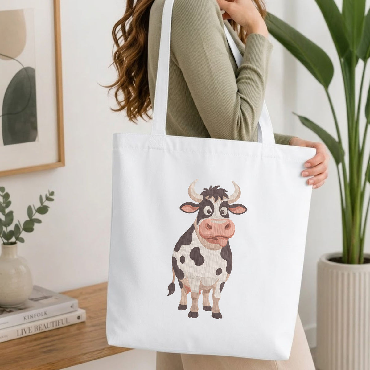 Woman carrying a white natural canvas tote bag featuring a funny cow machine embroidery design in a bright, modern interior setting