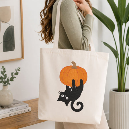 Woman carrying a white natural canvas tote bag featuring a mischievous black cat hanging upside down from a pumpkin machine embroidery design in a bright, modern interior setting