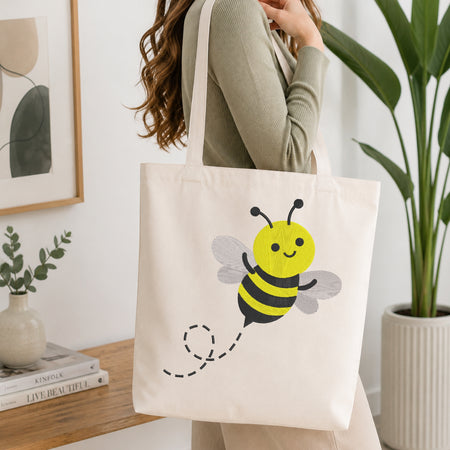 A woman wearing a sage green ribbed sweater carries a cream canvas tote bag decorated with a cute yellow and black bumblebee graphic. The scene is set in a bright, modern room with a wooden console table, a potted green plant, and a framed art print.