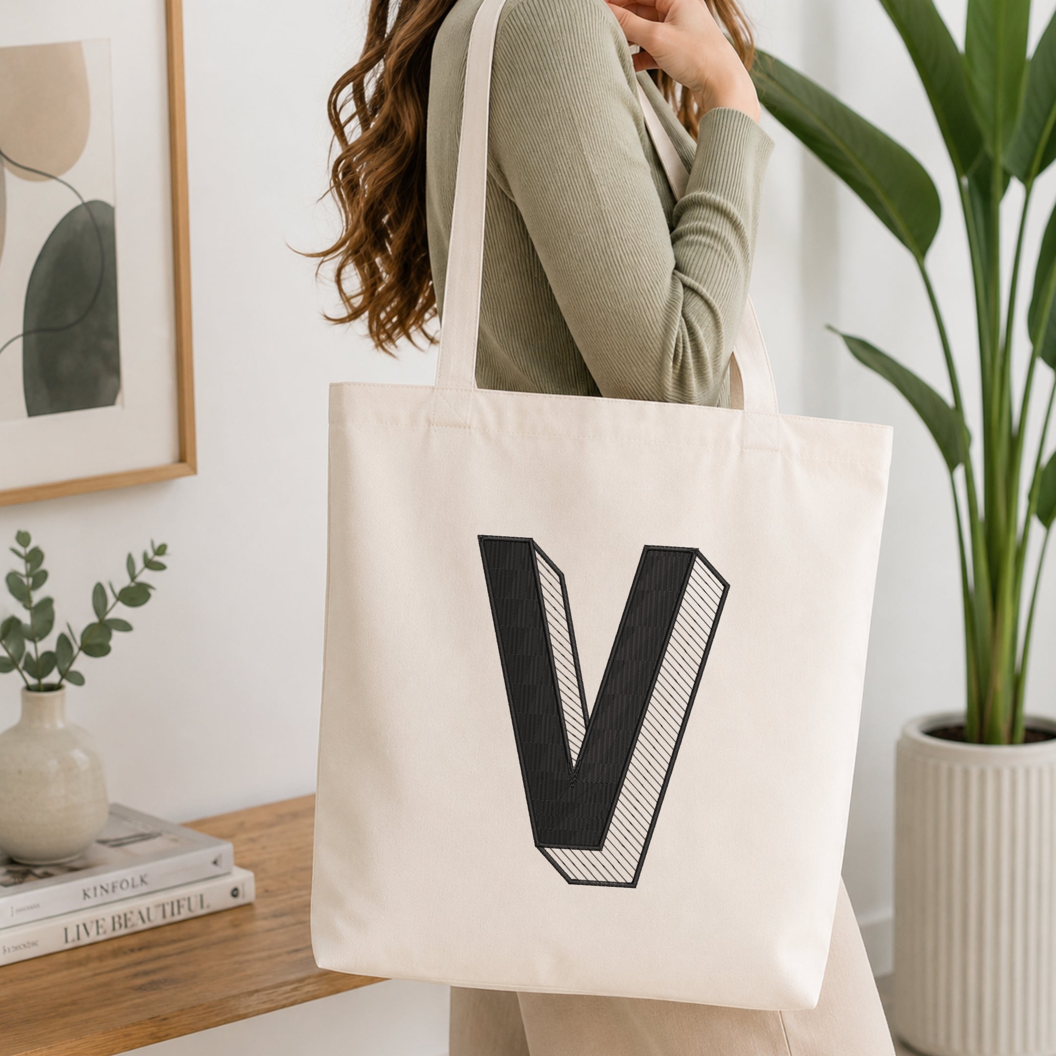 A person holding a natural canvas tote bag printed with an embroidered 3D block letter "V" design, set against a bright interior room background with green plants.