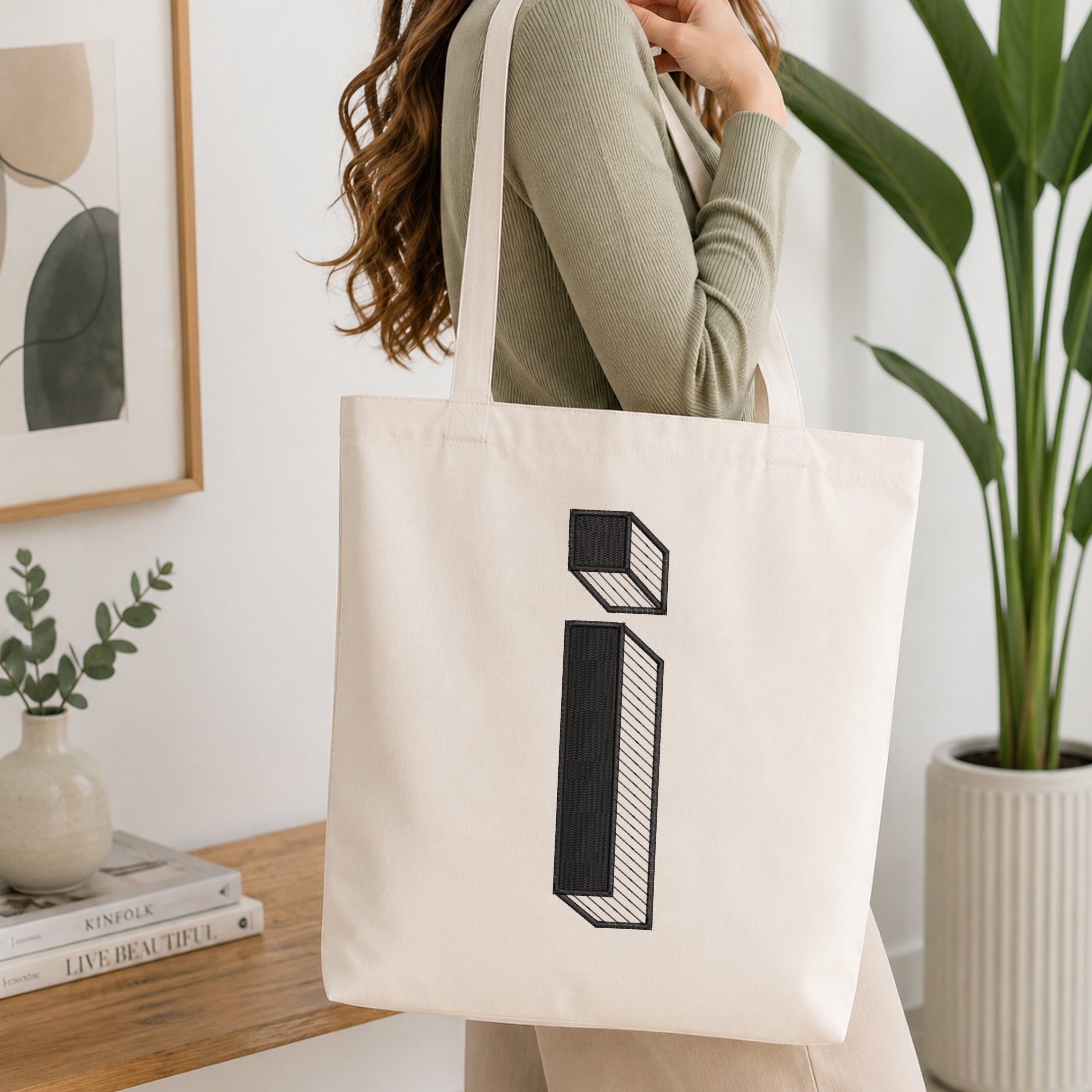 A woman wearing a sage green ribbed long-sleeve top carries a cream canvas tote bag with a large black geometric letter 'i' design on the front, set in a bright, modern room with a wooden table, framed art, and a potted plant.