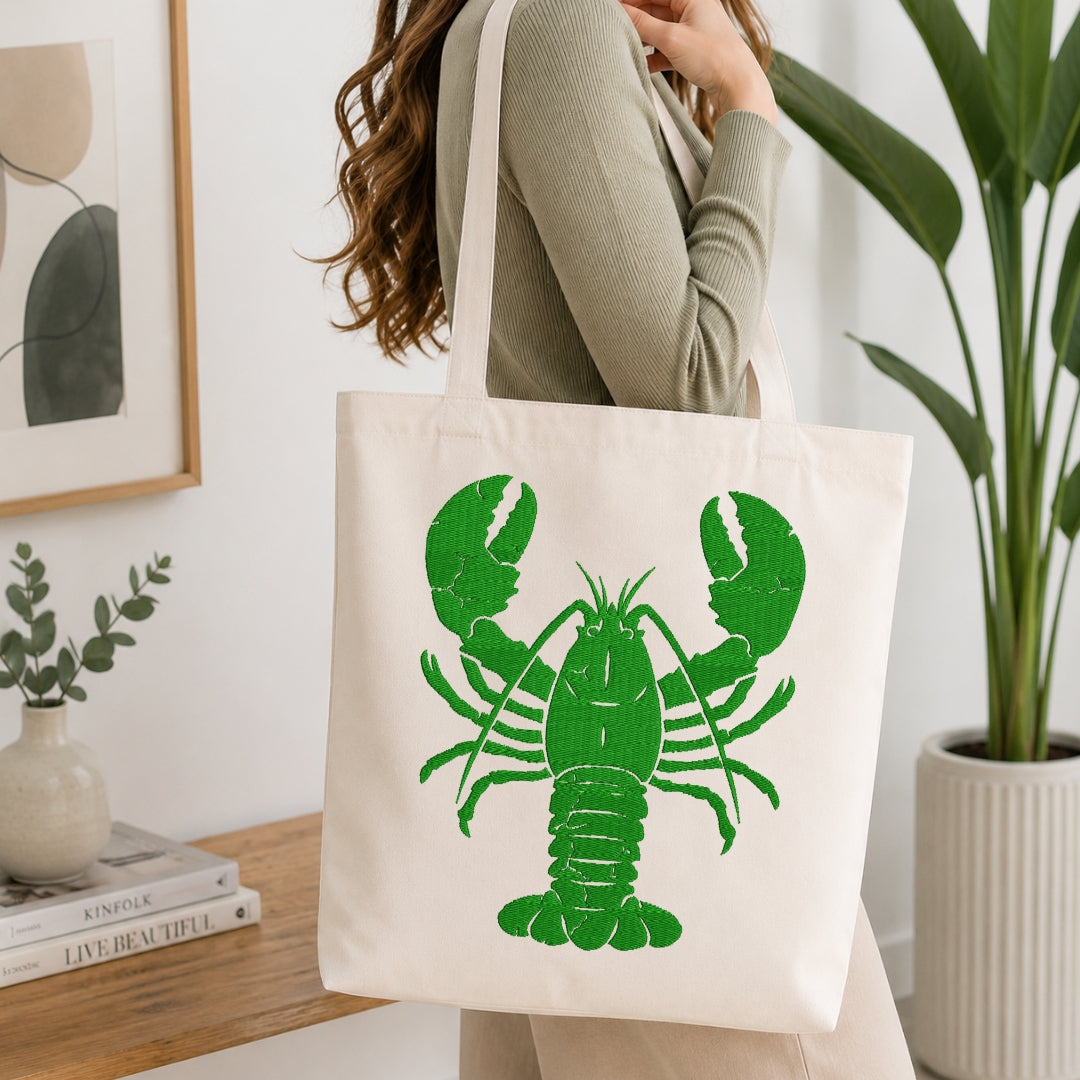 A person wearing a ribbed sage green top carries a cream canvas tote bag with a large green lobster graphic, standing in a bright, modern room with a wooden table, books, and a potted plant.