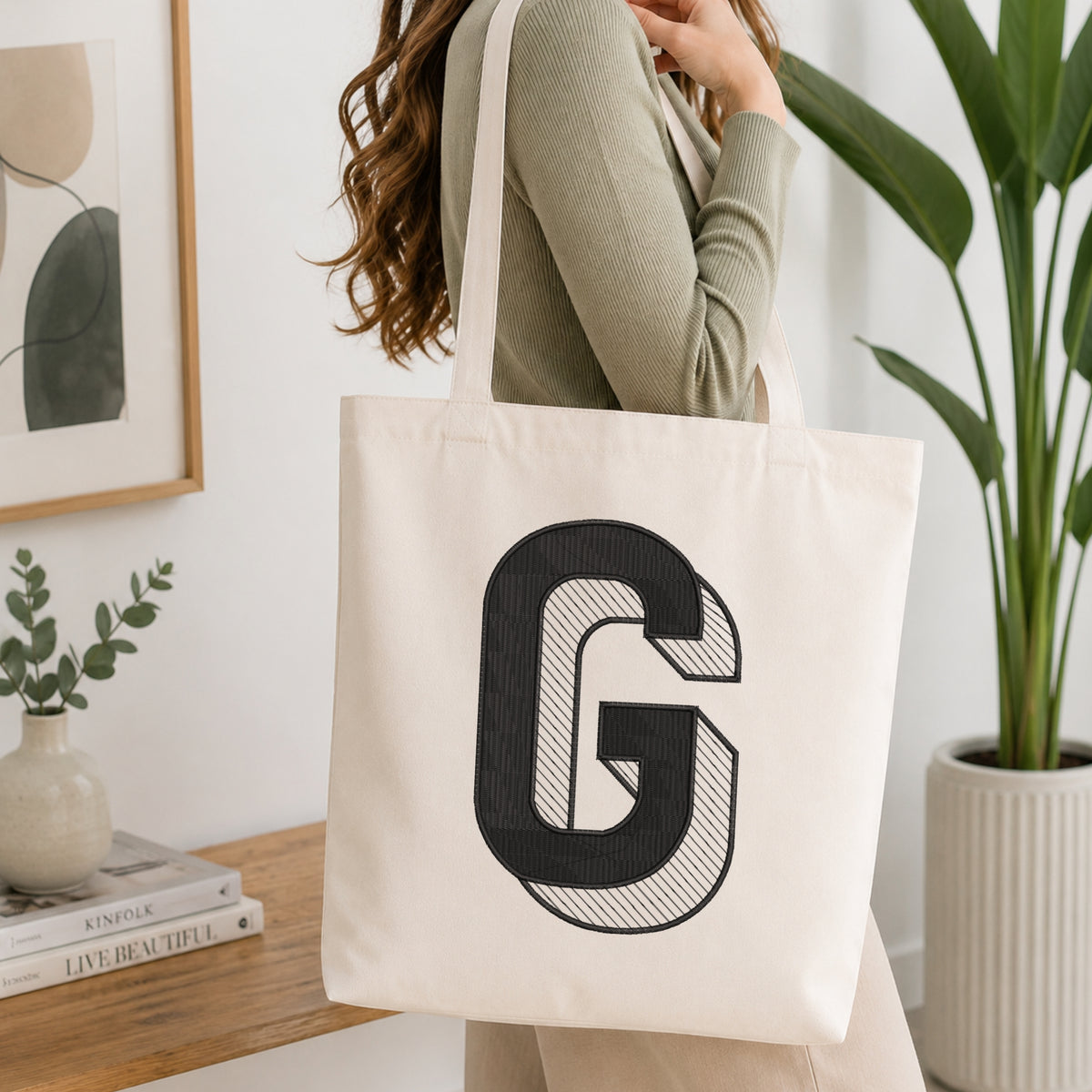 A woman wearing a sage green ribbed long-sleeve top carries a large off-white canvas tote bag with a bold black embroidered letter 'G' on the front, set in a bright, modern room with a wooden table, books, and a potted plant.