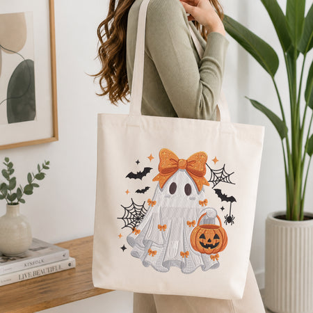 A person wearing a green long-sleeve top and carrying a natural canvas tote bag printed with the Halloween ghost with bow and pumpkin bucket machine embroidery design.
