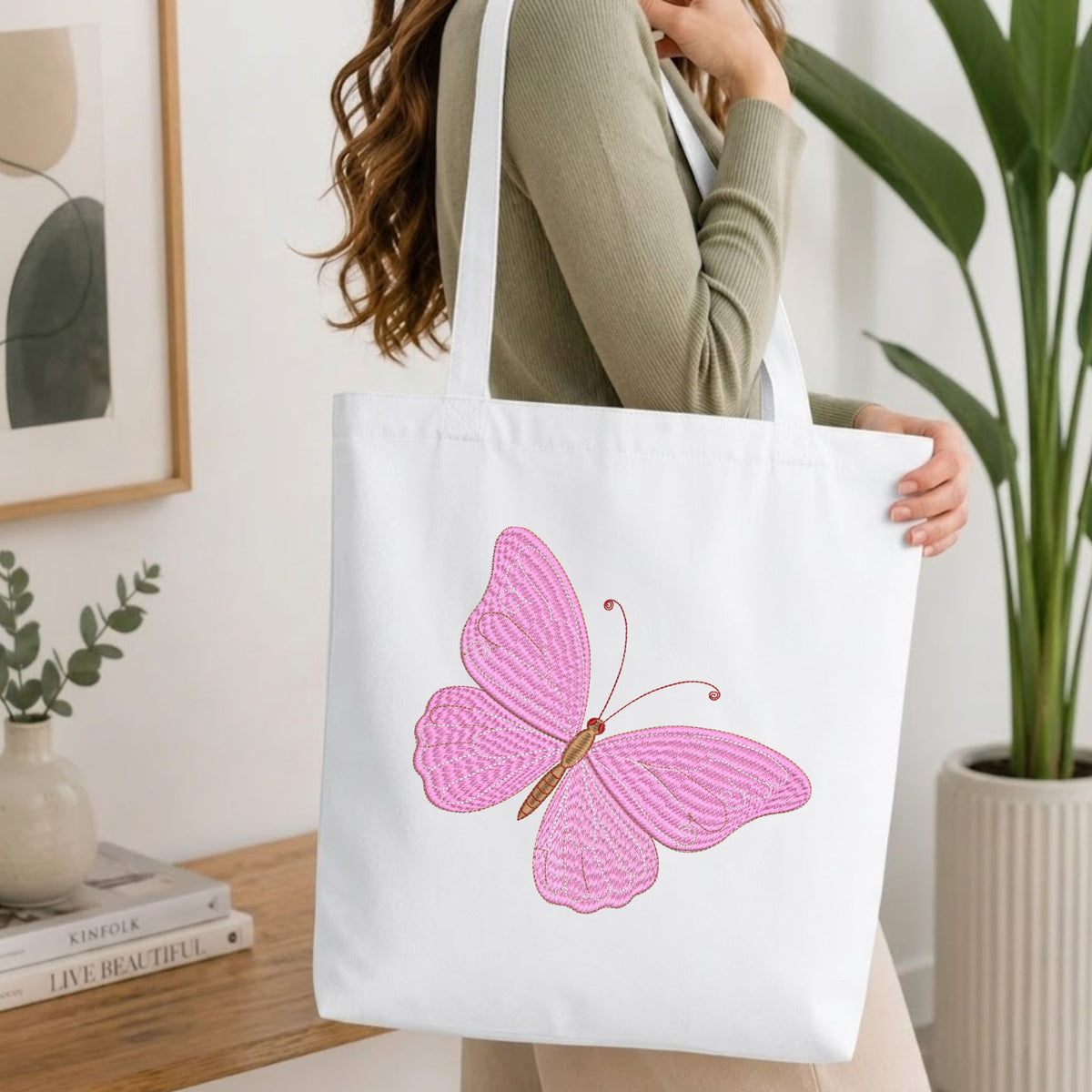 Woman carrying a white natural canvas tote bag featuring an embroidered elegant pink butterfly design in a bright, modern interior setting.