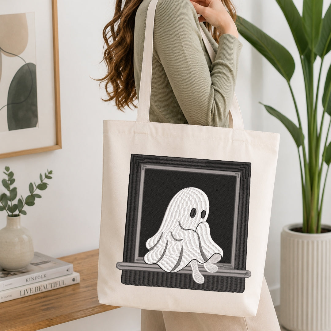 A woman wearing a sage green ribbed sweater carries a cream canvas tote bag with a black-framed graphic of a white wavy ghost sitting on a windowsill. The background shows a bright, modern room with a wooden table, books, a framed print, and a potted plant.
