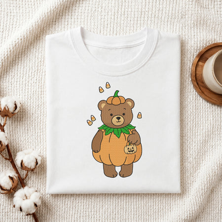 A folded white crewneck t-shirt featuring a cute cartoon graphic of a brown teddy bear dressed in a pumpkin costume, styled on a textured cream fabric with cotton bolls and a wooden cup in the background.