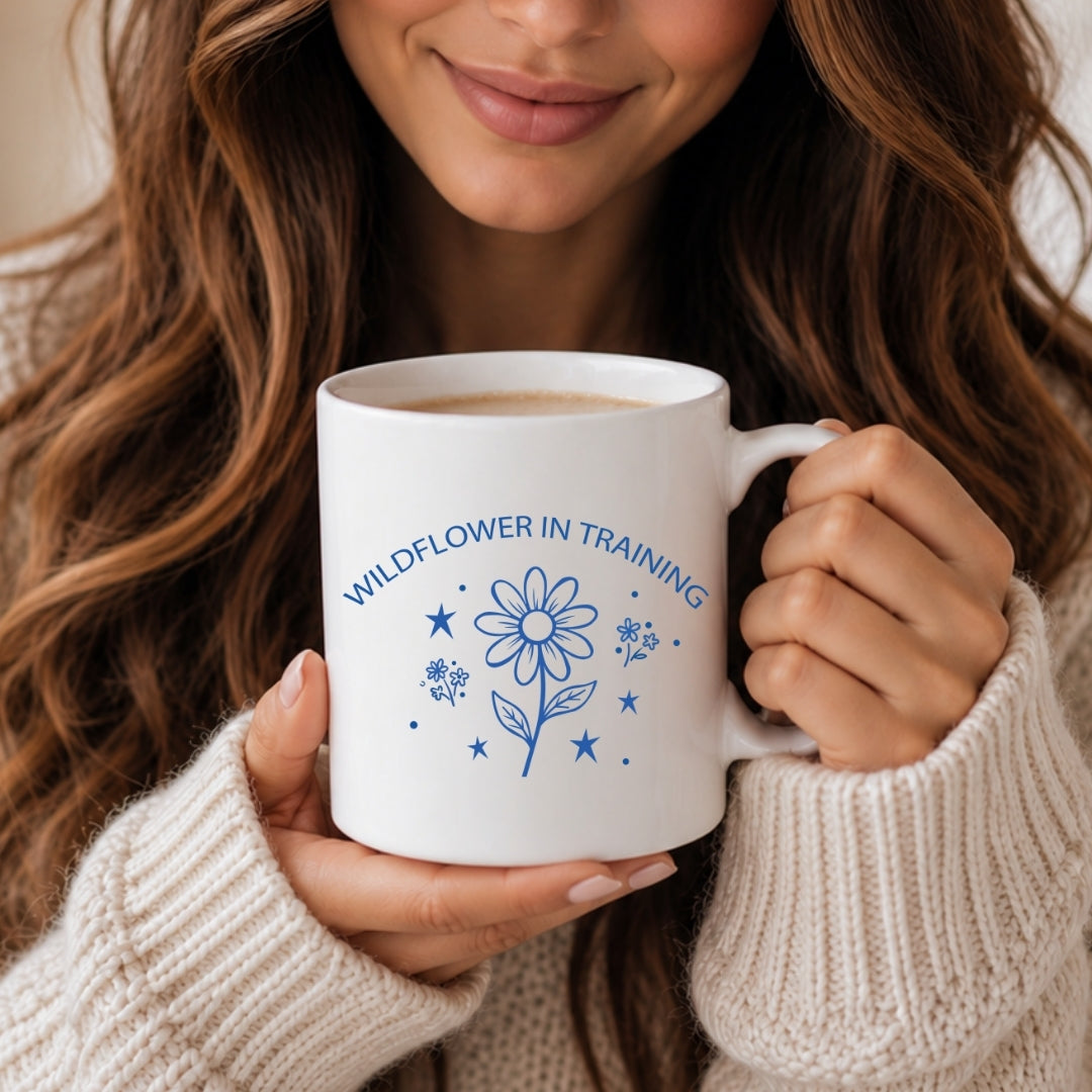 Woman with long wavy hair holding a white ceramic coffee mug printed with a blue minimal daisy "Wildflower in Training" graphic design.