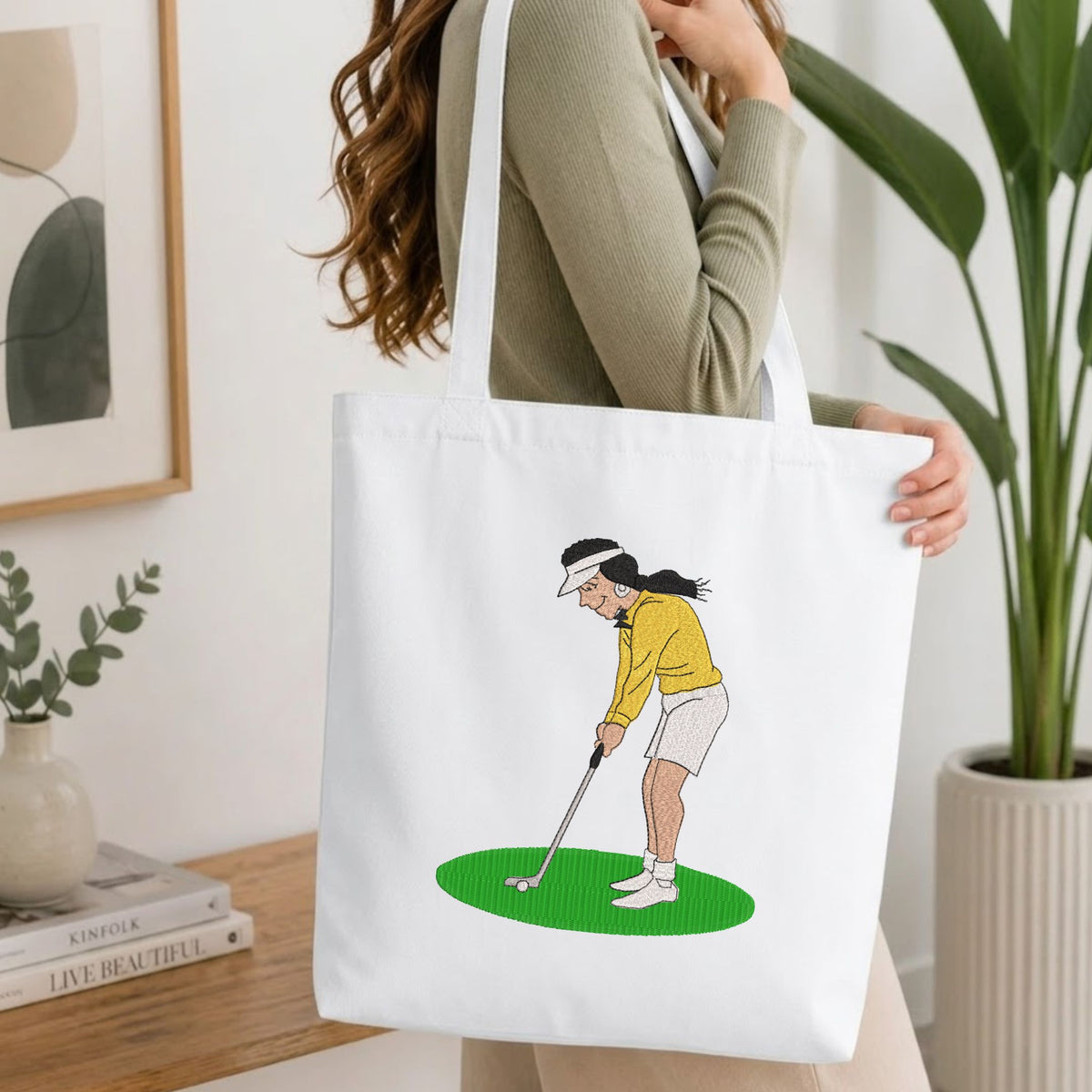 Woman carrying a white natural canvas tote bag featuring an embroidered woman playing golf design in a bright, modern interior setting   