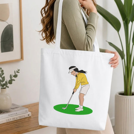 Woman carrying a white natural canvas tote bag featuring an embroidered woman playing golf design in a bright, modern interior setting   