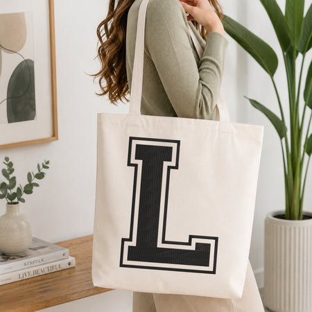 A woman wearing a sage green ribbed long-sleeve top carries a large off-white canvas tote bag with a bold black block letter 'L' outlined in white on the front. The scene is set in a bright, modern room with a wooden console table, a framed print, a small vase with greenery, and a potted plant.