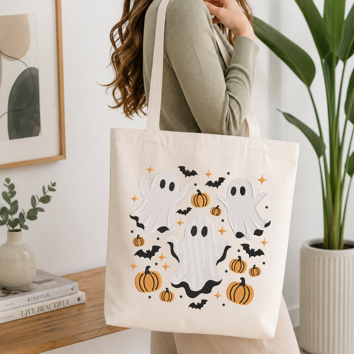 An off-white canvas tote bag with embroidered Halloween motifs including ghosts, pumpkins, bats, and stars, carried by a woman in a home setting.