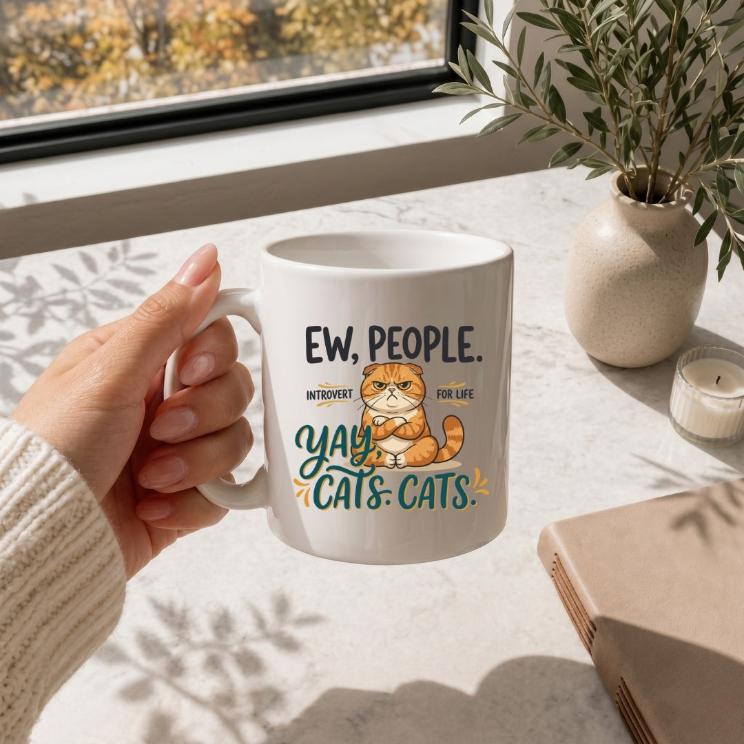 Hand holding a white ceramic coffee mug printed with a funny "Ew, People. Yay, Cats." grumpy cat graphic design on a sunlit marble tabletop.