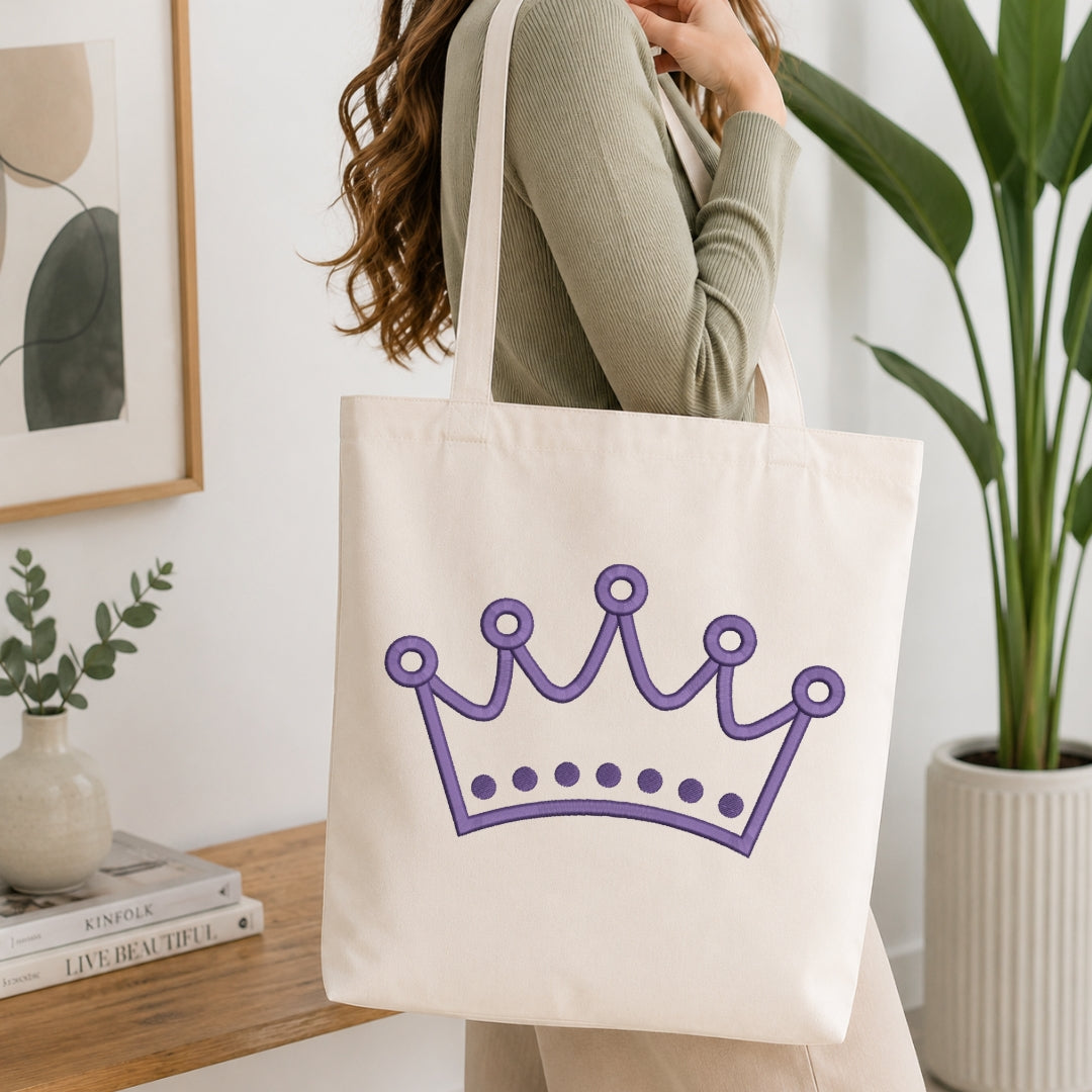 An off-white canvas tote bag with a purple crown graphic, carried by a woman wearing a sage green ribbed sweater in a bright, modern room with a wooden table and potted plants.