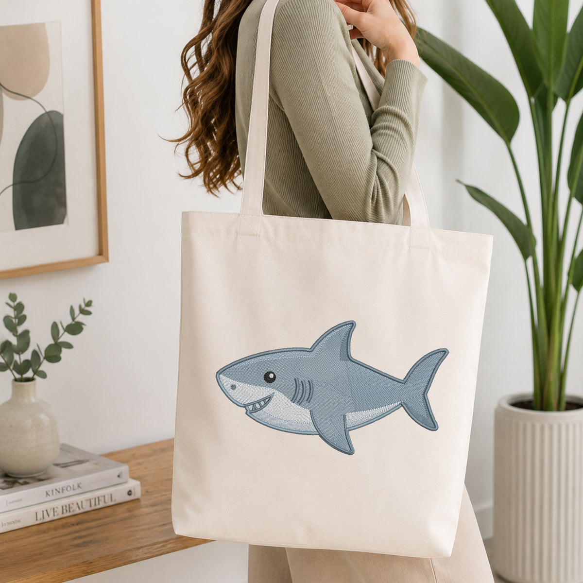 A woman wearing a sage green ribbed sweater carries a cream canvas tote bag with a blue-gray cartoon shark graphic on the front, set in a bright, modern room with a wooden table, books, and a potted plant.