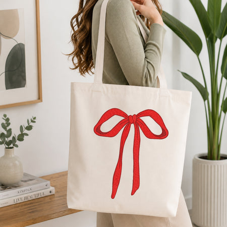 A woman wearing a sage green ribbed sweater carries a cream canvas tote bag with a large red bow graphic on the front, set in a bright, modern room with a wooden table and potted plants.