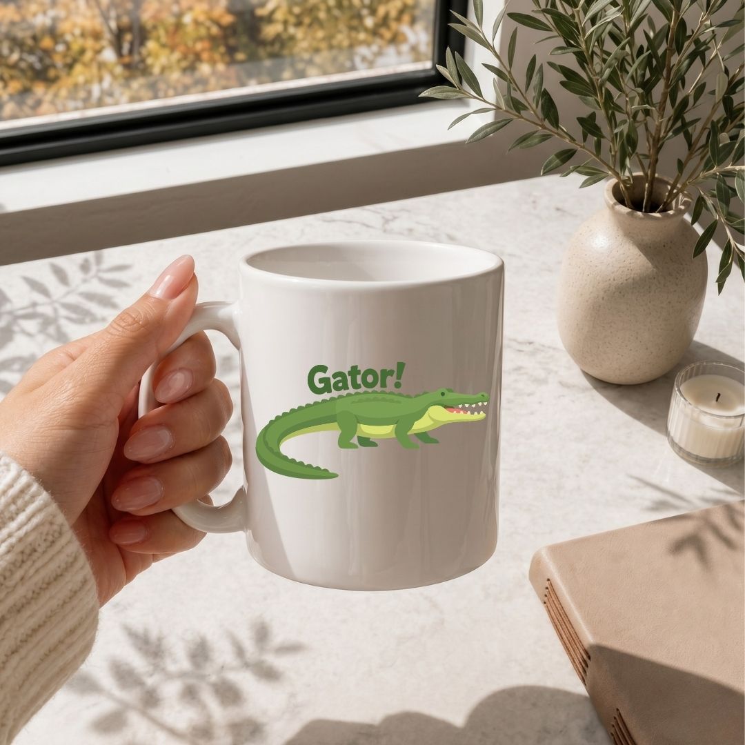 A person holding a white ceramic coffee mug by the handle near a sunny window, featuring a green alligator illustration with the text "Gator!".