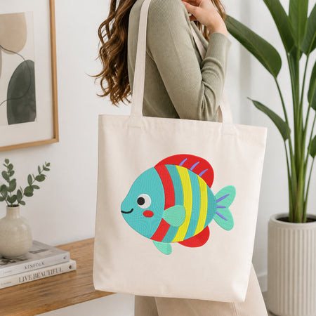 A woman wearing a sage green ribbed long-sleeve top carries a cream canvas tote bag with a colorful embroidered fish design. The background is a bright, modern room with a wooden side table, framed art, a small plant, and a large green potted plant.