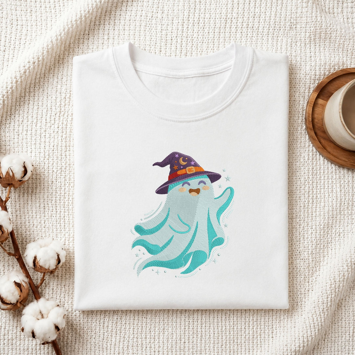 Folded white cotton t-shirt mockup featuring a blue witch ghost machine embroidery design, styled with cotton stems and a wooden cup   