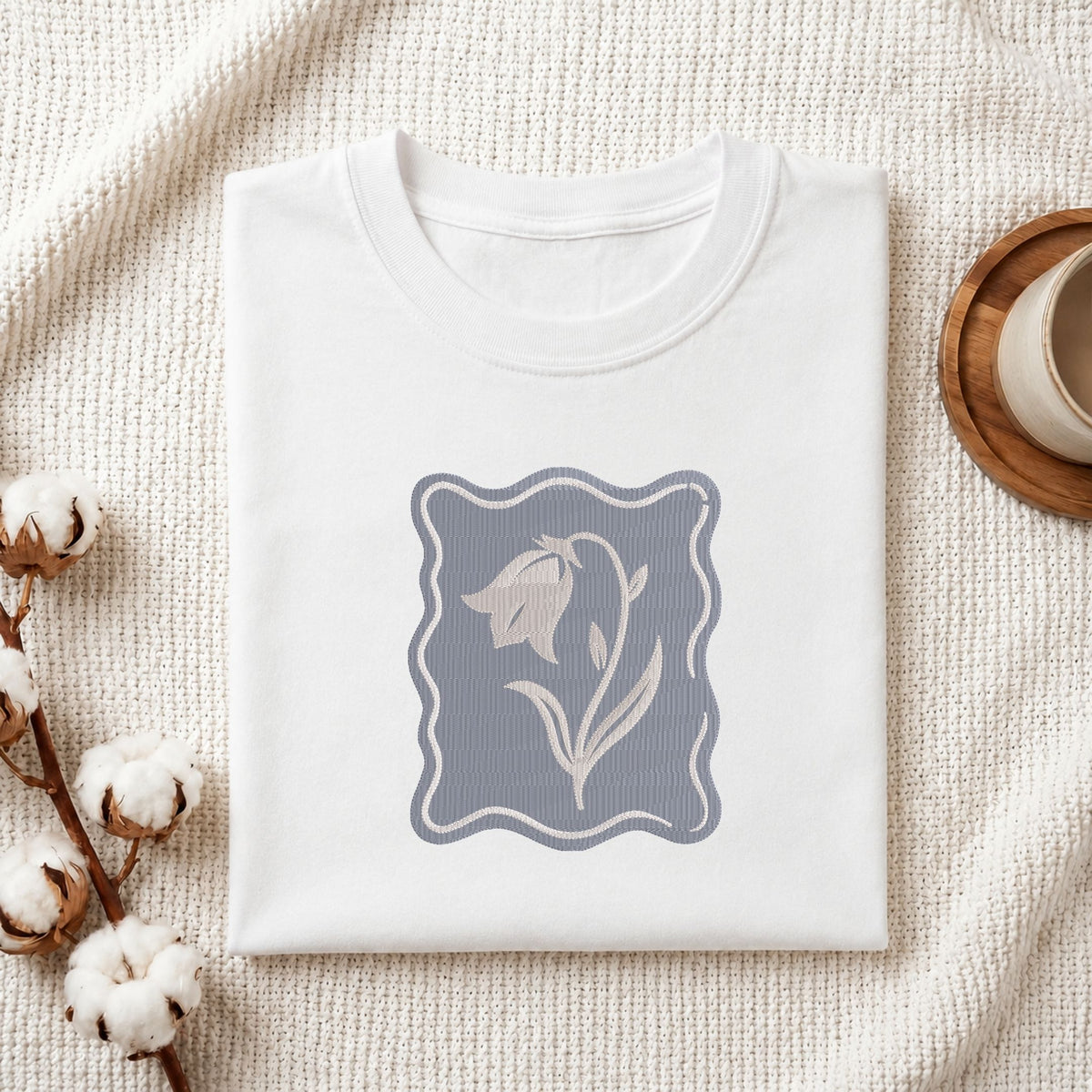 Folded white cotton t-shirt mockup featuring a minimalist bellflower machine embroidery design, styled with cotton stems and a wooden cup
