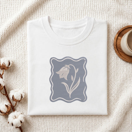 Folded white cotton t-shirt mockup featuring a minimalist bellflower machine embroidery design, styled with cotton stems and a wooden cup