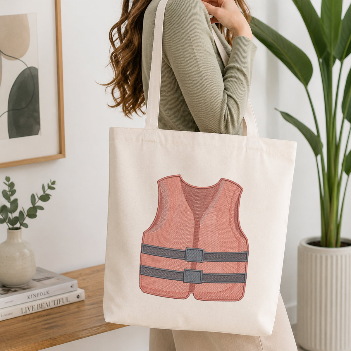 A woman wearing a sage green ribbed sweater carries a cream canvas tote bag featuring a printed illustration of a pink life vest with gray straps. The scene is set in a bright, modern room with a wooden table, framed art, a small plant, and a large green potted plant.