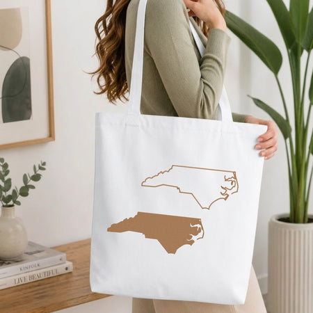 A woman wearing a sage green ribbed sweater carries a white canvas tote bag with a printed outline of North Carolina in tan and brown.