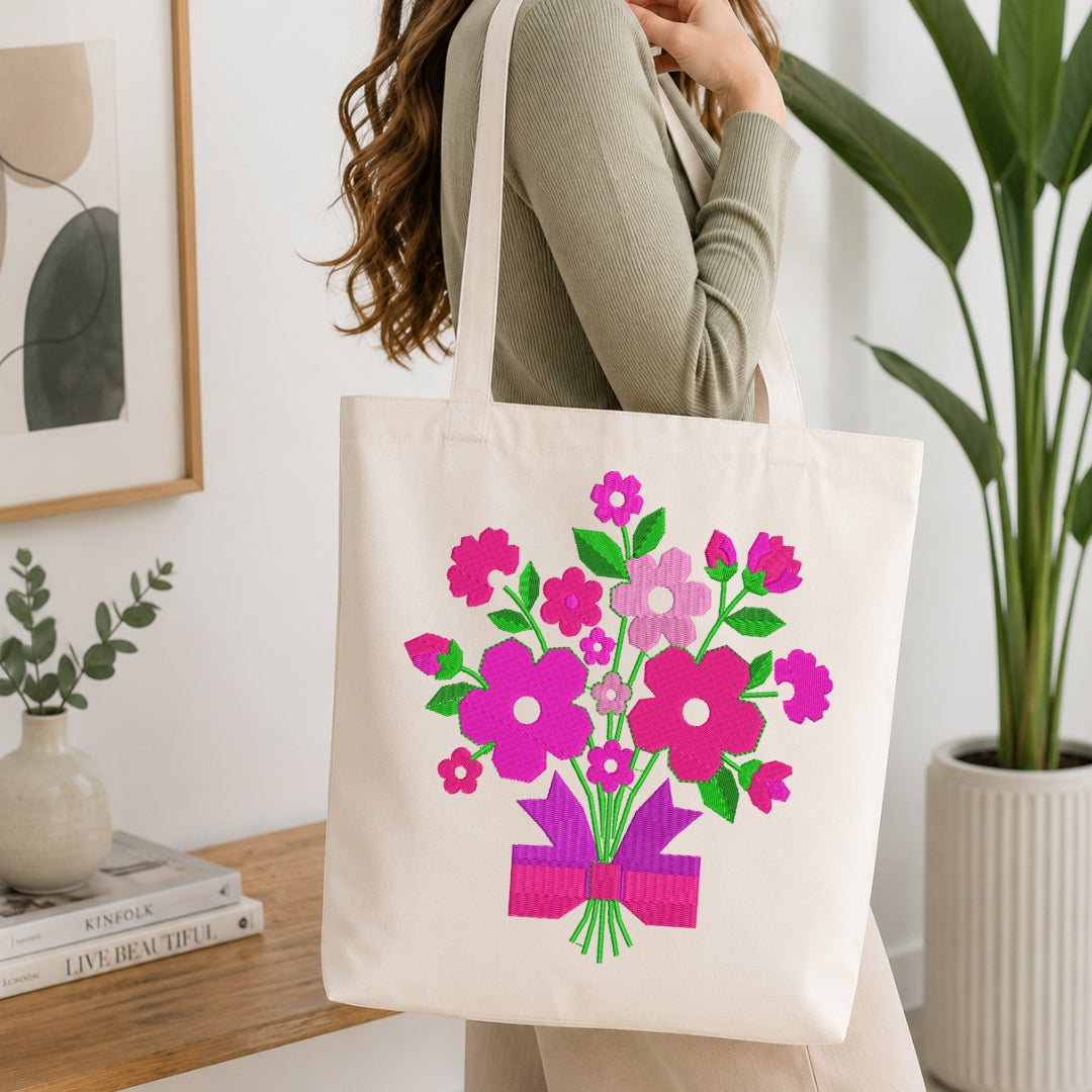 A woman wearing a sage green ribbed sweater carries a cream canvas tote bag decorated with a colorful floral bouquet print in pink, purple, and green, set against a bright, modern indoor background with a wooden table and potted plants.