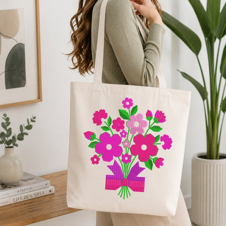 A woman wearing a sage green ribbed sweater carries a cream canvas tote bag decorated with a colorful floral bouquet print in pink, purple, and green, set against a bright, modern indoor background with a wooden table and potted plants.