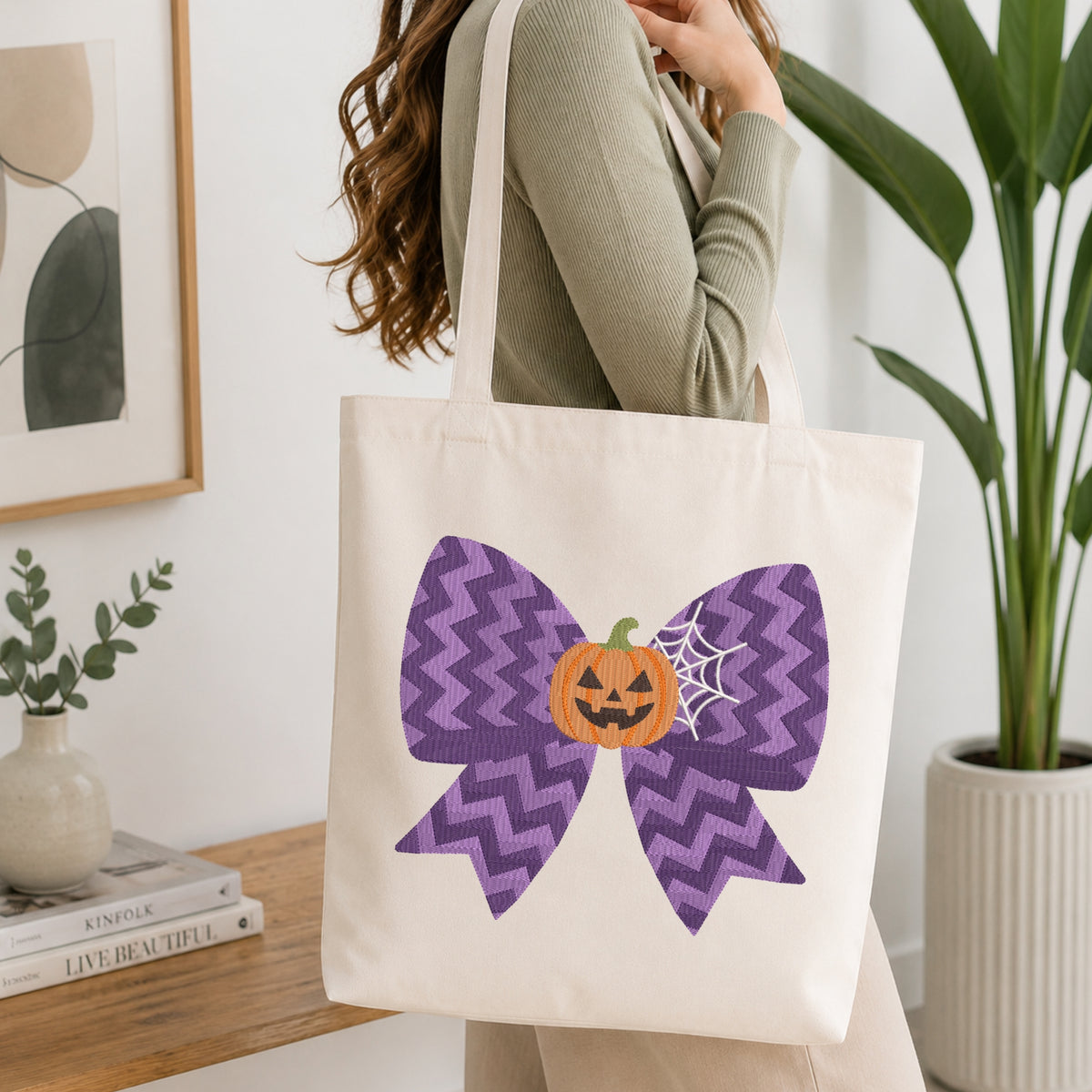 A person wearing a green long-sleeve top and carrying a natural canvas tote bag printed with the purple chevron Halloween bow machine embroidery graphic.