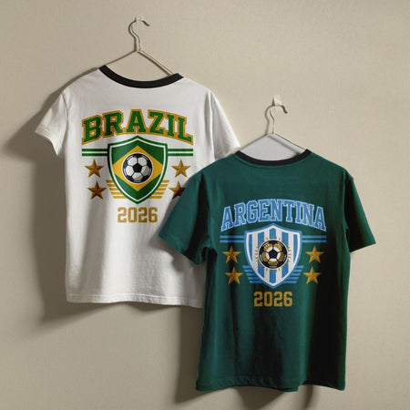 Two t-shirts, one white showing a green and gold Brazil soccer graphic and one dark green showing a light blue striped Argentina soccer design, hanging side by side on a wall.