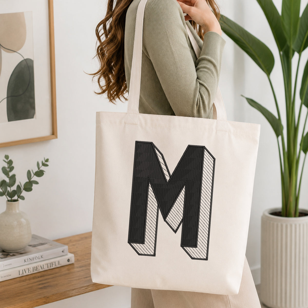 A woman wearing a sage green ribbed long-sleeve top carries a cream canvas tote bag with a large black block letter 'M' printed on the front, set in a bright, modern room with a wooden table, books, and a potted plant.