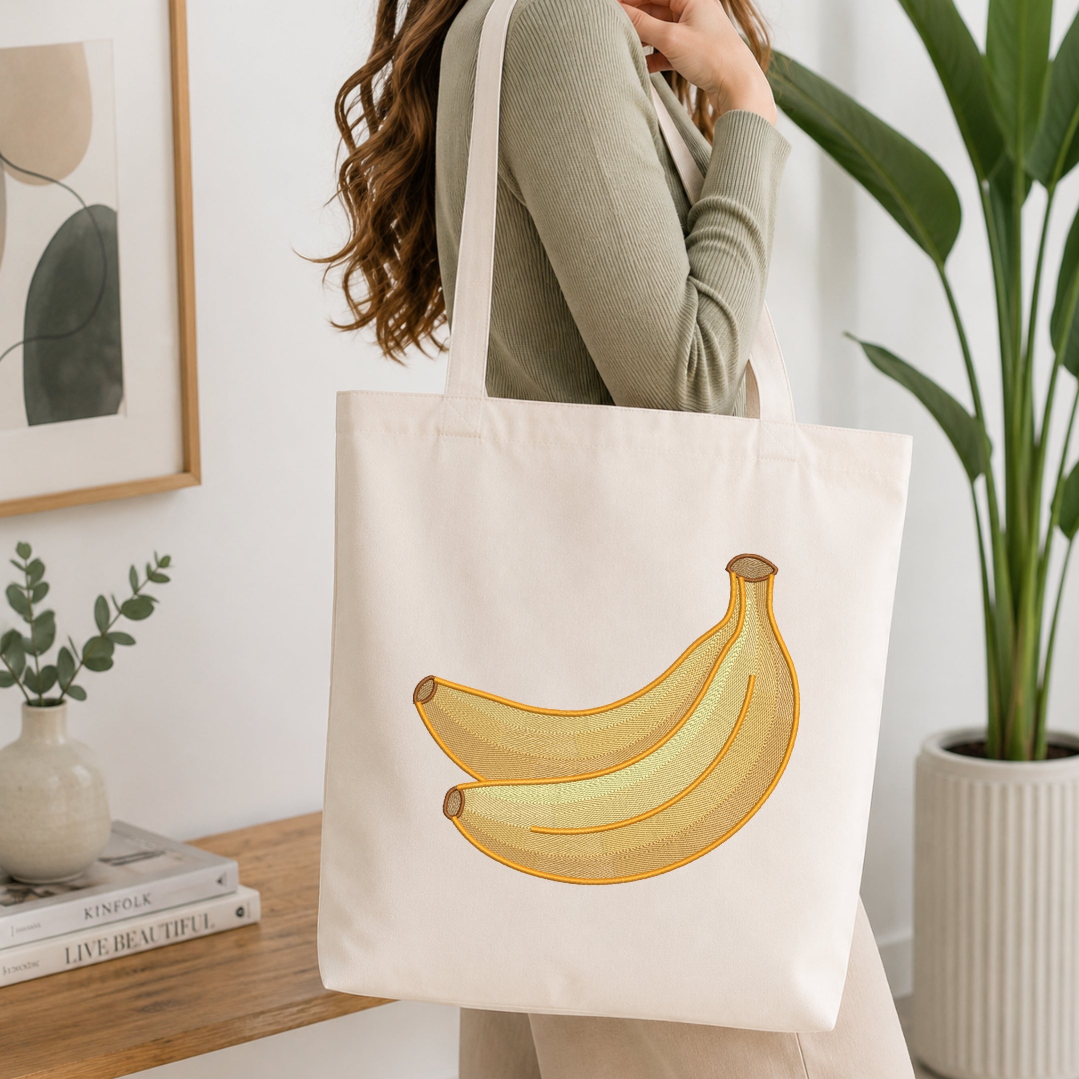 A woman wearing a sage green ribbed sweater carries a cream canvas tote bag with a yellow and gold banana graphic on the front, set in a bright, modern room with a wooden table, books, and a potted plant.