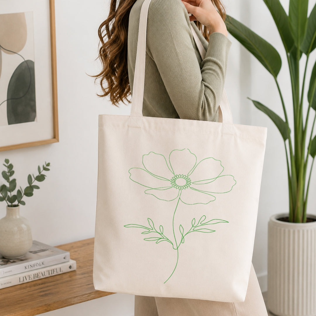 A woman wearing a sage green ribbed sweater carries a cream canvas tote bag with a green line-art flower design, set in a bright, modern room with a wooden table, books, and a potted plant.