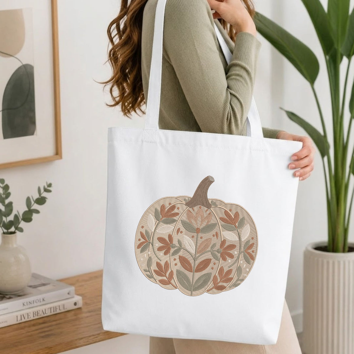 Woman carrying a white natural canvas tote bag featuring an embroidered floral pumpkin design in a bright, modern interior setting.
