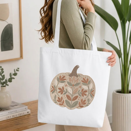 Woman carrying a white natural canvas tote bag featuring an embroidered floral pumpkin design in a bright, modern interior setting.