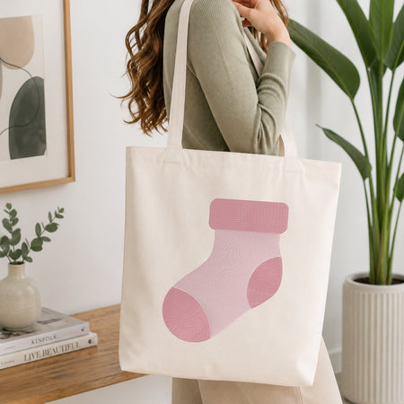 A woman wearing a sage green ribbed sweater carries a cream canvas tote bag with a colorful lollipop graphic. The background is a bright, modern room with a wooden shelf, framed art, and a potted plant.