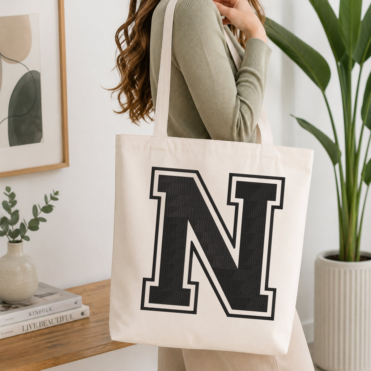 A woman wearing a sage green ribbed sweater carries a cream canvas tote bag with a large black block letter 'N' outlined in white on the front, set in a bright, modern room with a wooden console table and a potted plant.