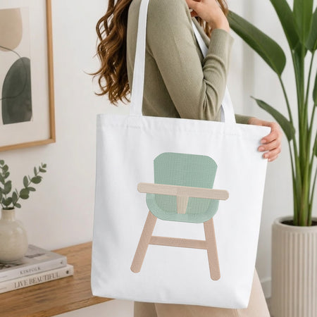 A woman wearing a sage green ribbed long-sleeve top carries a white canvas tote bag featuring a minimalist graphic of a chair with a mint green seat and beige wooden legs.