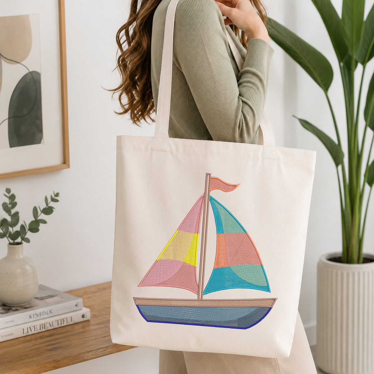 A woman wearing a sage green ribbed sweater carries a cream canvas tote bag with a colorful embroidered sailboat design. The background is a bright, modern room with a wooden shelf, framed art, and a potted plant.