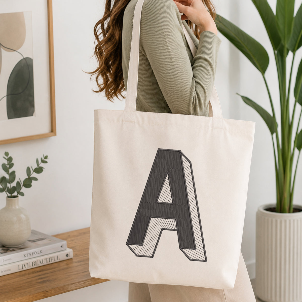 A woman wearing a sage green ribbed long-sleeve top carries a cream canvas tote bag with a large black block letter 'A' printed on the front, set in a bright, modern room with a wooden table, books, and a potted plant.