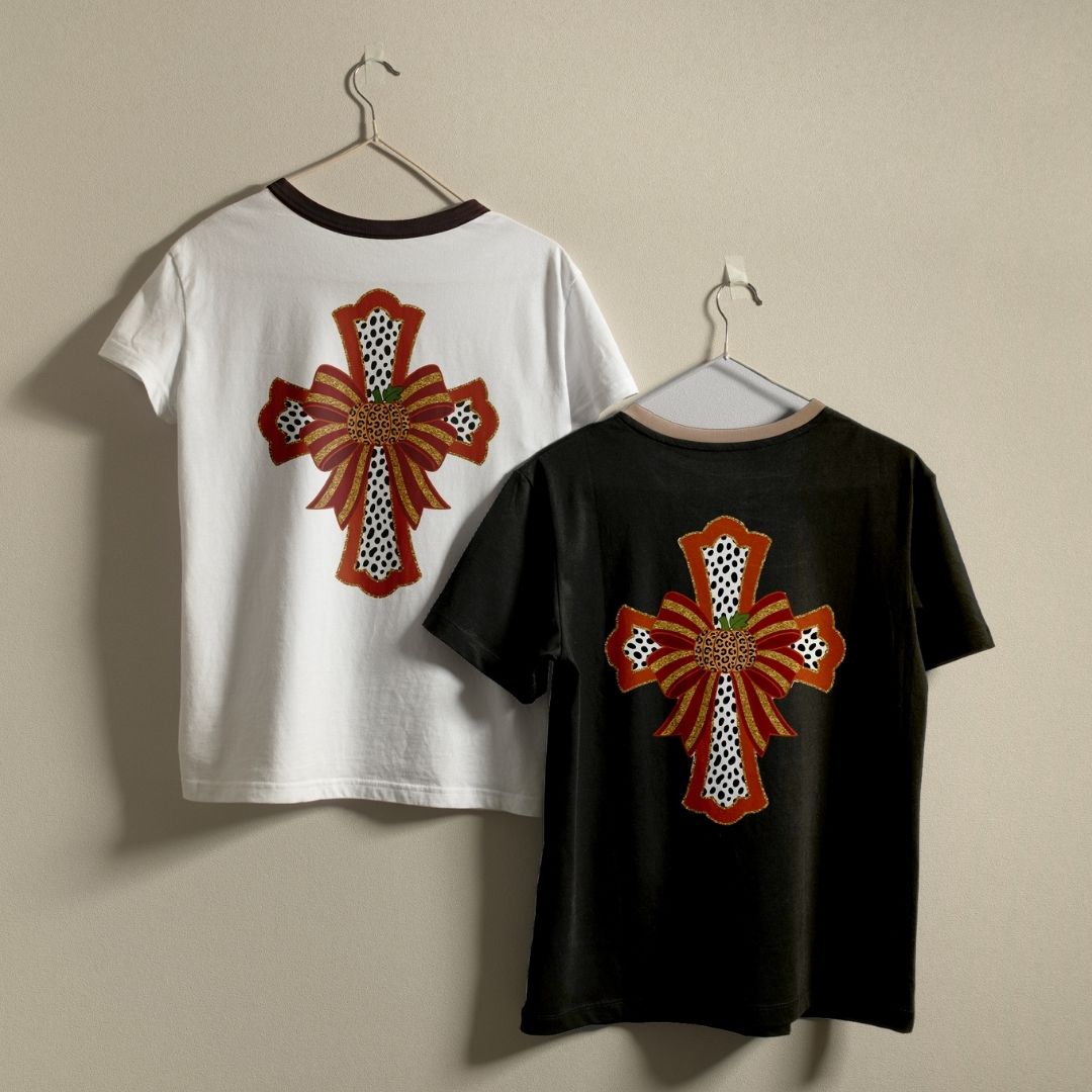 Two short-sleeve T-shirts hanging on hangers against a plain beige wall. The back of one white shirt and one black shirt both feature a large decorative cross graphic in rust red, black, white, and a small green accent.